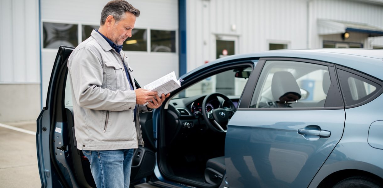 Un homme consulte le carnet d’entretien ouvert de sa voiture devant un garage, avec le tableau de bord et le compteur kilométrique visibles.