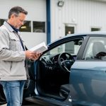 Un homme consulte le carnet d’entretien ouvert de sa voiture devant un garage, avec le tableau de bord et le compteur kilométrique visibles.