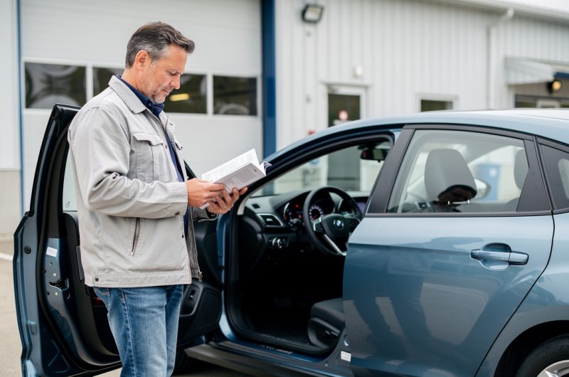 Un homme consulte le carnet d’entretien ouvert de sa voiture devant un garage, avec le tableau de bord et le compteur kilométrique visibles.
