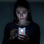 Une femme observe avec prudence un smartphone affichant une interface générique d’appel entrant, dans une ambiance sobre de cybersécurité.