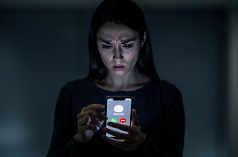 Une femme observe avec prudence un smartphone affichant une interface générique d’appel entrant, dans une ambiance sobre de cybersécurité.