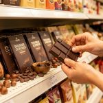 Main d’une femme comparant des tablettes de chocolat sans marque dans un rayon, entourées de cabosses de cacao, de fèves et de noisettes.